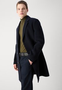 Marineblauer Wollmantel mit Reverskragen, drei Knöpfen und lockerer Passform, kombiniert mit einem olivgrünen Rollkragenpullover und dunklen Hosen.