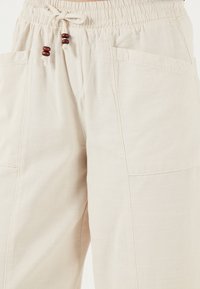 Beige wide-leg trousers made of textured fabric. Features an elastic waistband with drawstrings and wooden accents, plus two side pockets.