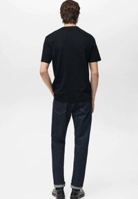 Bărbat îmbrăcat cu un tricou simplu negru cu mânecă scurtă, blugi bleumarin cu manșete și pantofi negri, stând cu mâinile în spate, cu fața întoarsă.