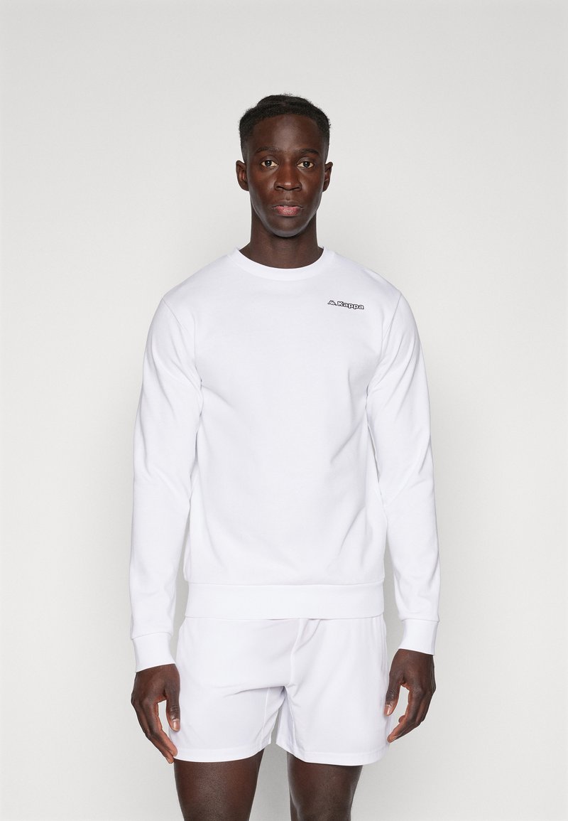 Sweatshirt blanc à col rond avec manches longues et poignets côtelés, doté d'un petit logo noir sur la poitrine gauche, associé à un short blanc.