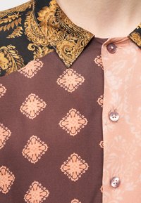 Πολύχρωμο πουκάμισο με μαύρο και χρυσό σχέδιο στο γιακά, ροζ και καφέ ύφασμα με λουλουδάτα μοτίβα και ροζ κουμπιά.