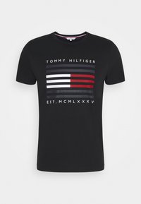 Černé bavlněné tričko s grafikou, která má horizontální pruhy v červené, bílé a šedé barvě, a textem "TOMMY HILFIGER" a "EST. MCMLXXXV."