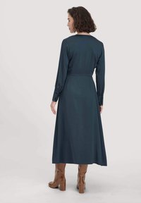 Mekko, jonka hihat ovat pitkät ja väri tumman teal, vyötärö on istuva ja hameosa laskeutuu ilmavasti. Pinta on sileä, takana on kauniit kaula-aukko yksityiskohdat, ja asusteena on ruskeat polvimittaiset saappaat.
