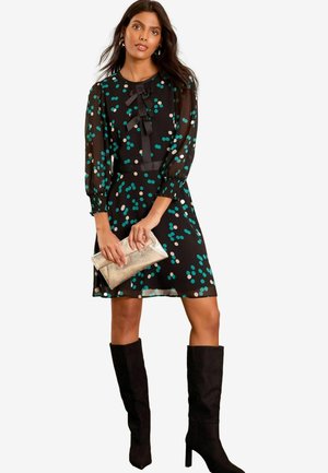 Schwarzes Kleid mit kurzen Puffärmeln, Teal- und Beigetönen im Punktmuster, zwei Schleifen am Hals, kombiniert mit einer metallischen Clutch und knielangen schwarzen Stiefeln.