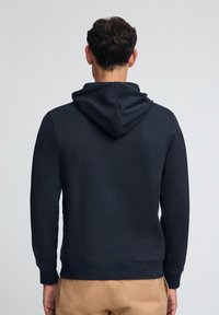 Mörkblå huva av mjukt tyg, med dragsko i huvan, ribbade ärmslut och en enkel design utan synliga logotyper eller mönster.