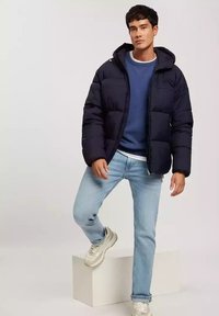 Granatowa kurtka puchowa z kapturem, posiadająca pikowany wzór i zamek błyskawiczny na przodzie, w połączeniu z jasnoniebieskim swetrem i jasnymi dżinsami.