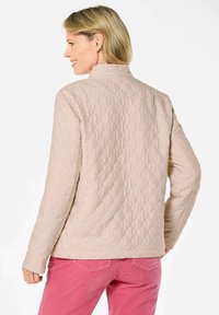 Beige gewatteerde jas met een hoge kraag en lange mouwen. Kenmerkt zich door ruitpatronen en gestikte naden. Draagt met roze broeken.