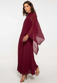 Abito lungo semi-trasparente color bordeaux con un design fluido, caratterizzato da un collo alto e maniche ampie, abbinato a tacchi dorati con lacci.