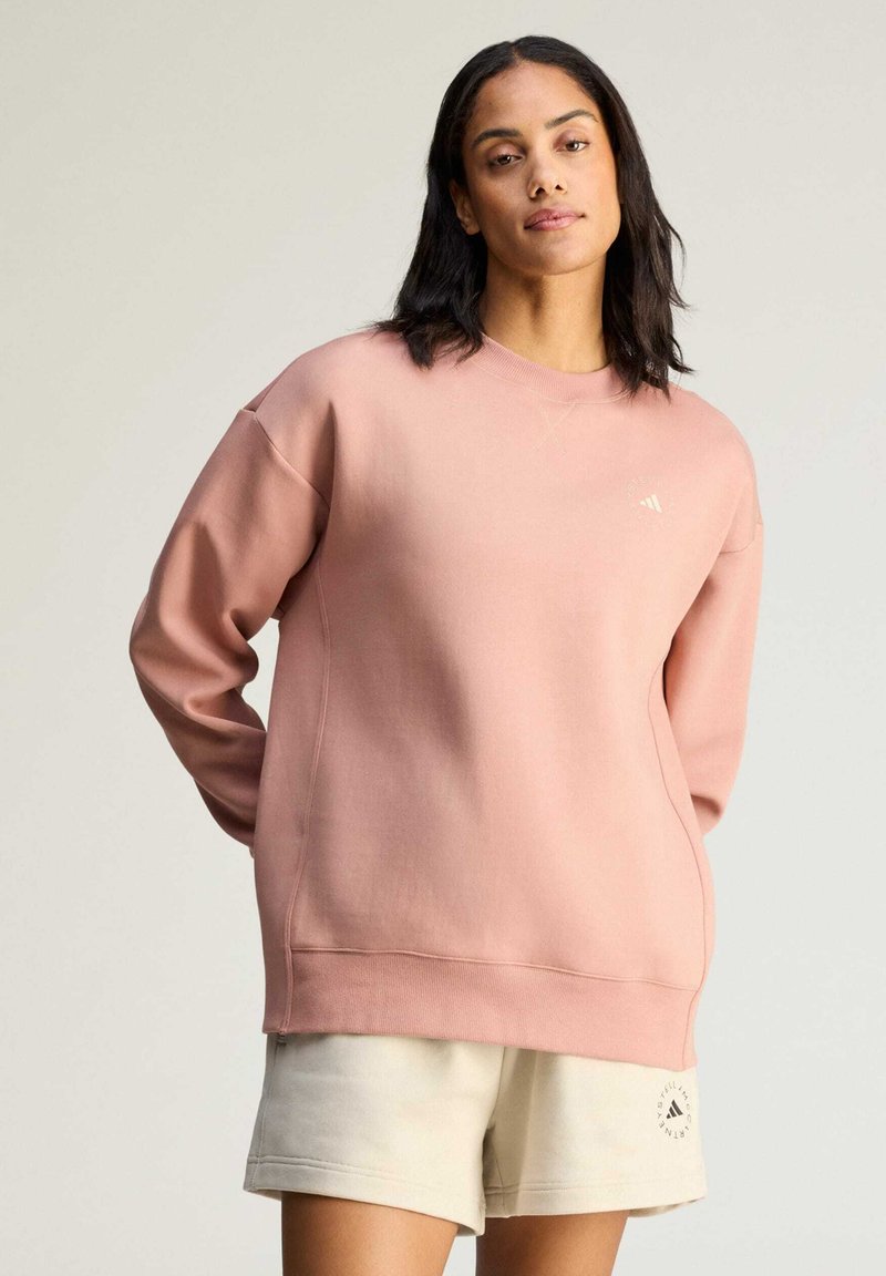 Kvinde stående med hænderne bag ryggen iført oversize støvet lyserød sweatshirt og lyse beige shorts mod en ensfarvet baggrund.
