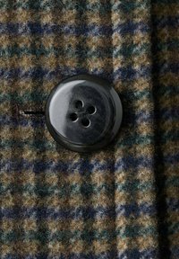 Wollen stof in een bruin, groen en blauwe houndstooth patroon, met een grote zwarte viergaatjesknop. Zachte textuur met een lichte fuzzy uitstraling.