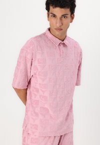 Pink polo shirt featuring a textured pattern of square logos, with a collared neckline and short sleeves. Soft, fluffy fabric with intricate detail.