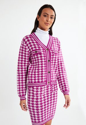 Set din cardigan și fustă tricotate, cu un model houndstooth roz și alb, având decolteu adânc în V, nasturi și două buzunare frontale.
