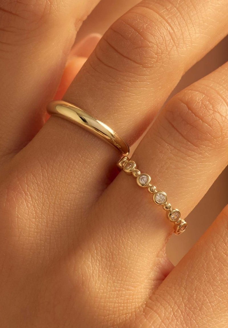 Bague en or avec une surface lisse et polie à côté d'une bande en or comportant cinq pierres claires incrustées, sur une main humaine.