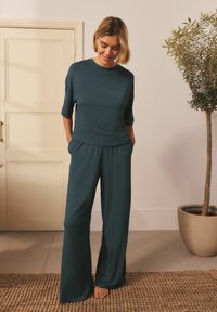 Tumši zilgani loungewear komplekts, kas sastāv no īsām piedurknēm t-krekla un platām biksēm, izgatavots no mīksta auduma ar brīvu piegulšanu. Redzami vienkārši dizaina elementi.