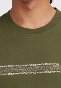 Olijfgroene katoenen t-shirt met een horizontaal crèmekleurig tribaal patroon over de borst. Ronde halslijn en korte mouwen.