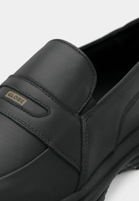 Gros plan sur une chaussure noire en cuir sans lacets avec une semelle texturée et une petite étiquette dorée marquée Globe sur la bride supérieure.