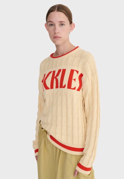 Jeune femme portant un pull en tricot beige avec l'inscription rouge "KKLES" et un bord rouge assorti, associée à un pantalon ample kaki, debout devant un fond blanc.