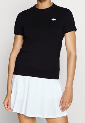 Frau trägt ein schwarzes kurzärmeliges Shirt mit weißem Logo und einem weißen plissierten Rock, steht vor einem einfarbigen hellen Hintergrund.