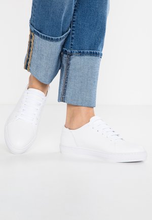 Person iført opbukte blå jeans og simple hvide snøresneakers, stående på en hvid overflade foran en neutral baggrund.