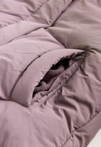 Prošívaná deka z jemného mauve materiálu, která obsahuje výraznou kapsu odhalující tmavě šedou fleecovou podšívku, s hladkou texturou a pečlivě šitými švy.