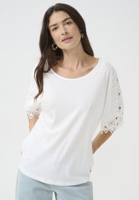 Hvid kortærmet t-shirt med blonderdetaljer på ærmerne; blødt stof; afslappet pasform; rund halsudskæring; båret med lyseblå jeans.