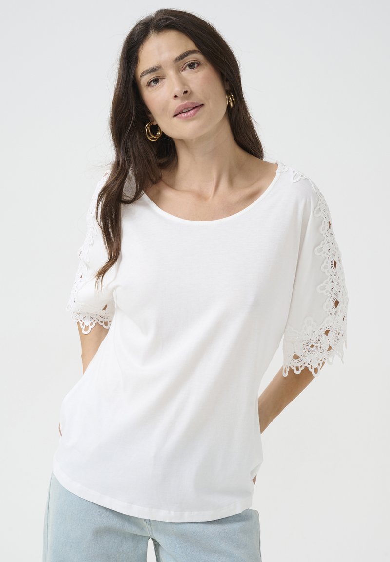 Hvid kortærmet t-shirt med blonderdetaljer på ærmerne; blødt stof; afslappet pasform; rund halsudskæring; båret med lyseblå jeans.