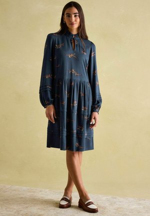 Teal Kleid mit langen Ärmeln, gerüschtem Rock, floralem Muster, Schlüsselaugen-Ausschnitt und gestreiften Manschetten, kombiniert mit braunen und weißen Schuhen.