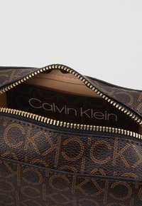 Borsa Calvin Klein con un esterno in pelle texturizzata marrone scuro, caratterizzata da un motivo a logo ripetuto in marrone chiaro e una chiusura con zip dorata.