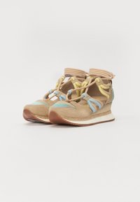 Design di scarpa con una tomaia in suede beige, elastici blu e gialli, punta rotonda e una suola bianca spessa con un battistrada marrone testurizzato.