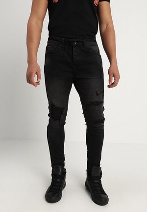 Man wearing black ripped skinny jeans, black high-top sneakers, and a black t-shirt with orange designs, standing against a plain background.