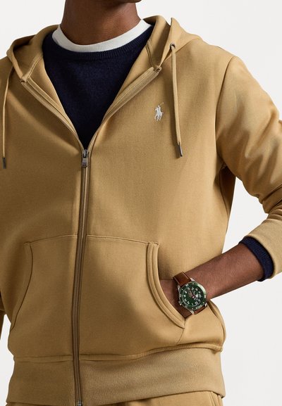 Sweatshirt bege com fecho, com um bolso frontal e detalhes em prata, sobre um suéter azul escuro, combinado com um relógio de pulso castanho com mostrador verde.