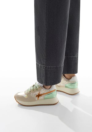 Sneakers feature a cream upper with glitter accents, green and orange highlights, flat laces, and a textured sole in a light brown color.