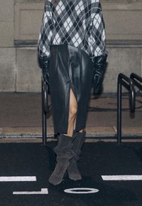 Pull surdimensionné en losanges noir et blanc, jupe midi en cuir noir texturé avec fente latérale, et bottes molletonnées grises en daim jusqu'au genou.