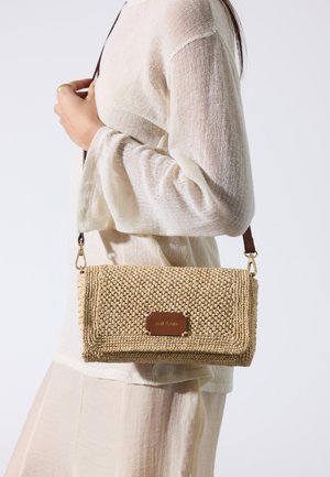 Mujer con suéter crema y falda sosteniendo un bolso cruzado beige texturizado tejido con una correa marrón y placa con logo.