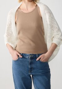 Brązowy prążkowany top bez rękawów nałożony pod białym dzianinowym kardiganem; niebieskie jeansy z wysokim stanem z przednimi kieszeniami. Teksturowana tkanina i swobodny krój.