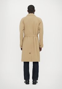Trench coat bej cu talie încinsă, manșete cu nasturi și fante la spate, purtat peste blugi negri și pantofi negri.