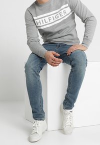 Uomo seduto su un blocco bianco, indossa una felpa grigia Hilfiger, jeans blu e sneakers bianche, con le mani appoggiate sulle gambe.