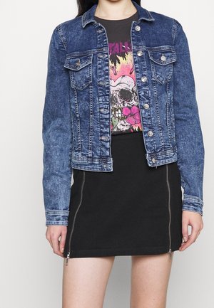 Kvinde iført blå denimjakke over sort T-shirt med grafisk kraniummotiv og sort nederdel med lynlåse foran.