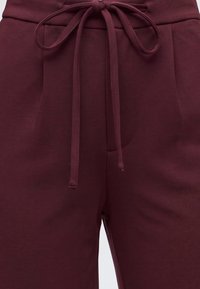 Gros plan sur un pantalon bordeaux avec un cordon de serrage en tissu noué à la taille et des détails cousus près des poches avant.