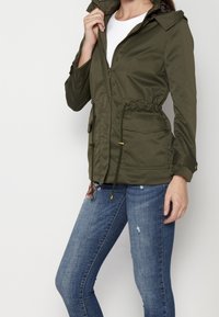 Kobieta w oliwkowej kurtce z przednimi kieszeniami i sznurkiem, nosząca niebieskie, przetarte dżinsy z haftem kwiatowym na górnej części uda.