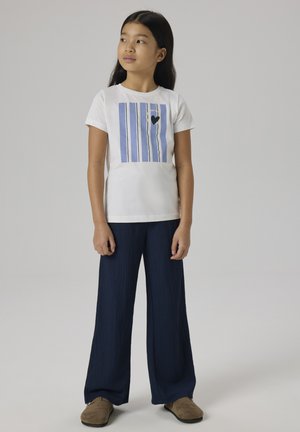 Fille aux longs cheveux noirs portant un t-shirt blanc avec des rayures verticales bleues et un cœur, un pantalon large bleu marine et des chaussures marron, debout regardant vers la gauche.