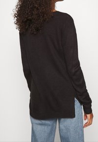 Maglione nero lavorato a maglia con maniche lunghe, fessure laterali e vestibilità rilassata. Il materiale sembra morbido con una superficie liscia. Abbinato a jeans azzurri chiari.