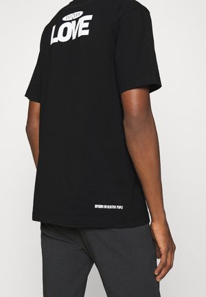 Personne portant un T-shirt noir avec le texte blanc "DRYKORN LOVE" sur le haut du dos et "DRYKORN FOR BEAUTIFUL PEOPLE" près de l'ourlet inférieur.