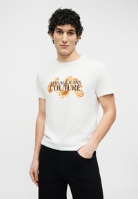 Νέος άνδρας με σγουρά μαλλιά, που φοράει λευκό t-shirt Versace Jeans Couture με πορτοκαλί διακοσμητικό σχέδιο και μαύρο παντελόνι.