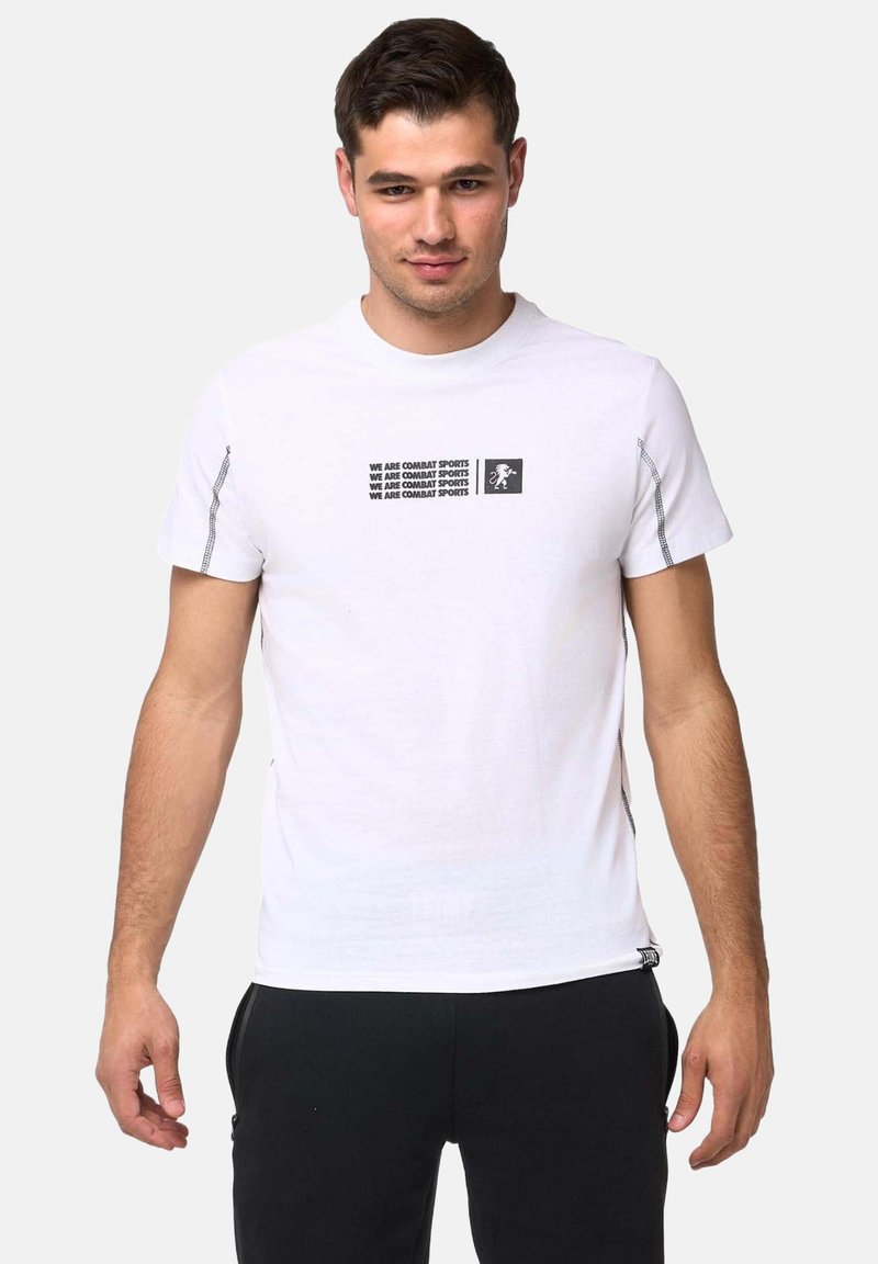 Homme portant un t-shirt blanc avec le texte « WE ARE COMBAT SPORTS » et un petit logo de lion, associé à un pantalon noir, debout sur un fond blanc.