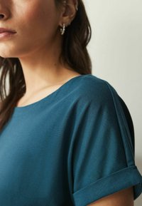 Tyrkysový top s krátkým rukávem a kulatým výstřihem, s ohrnutými manžetami; měkká textilie je viditelná s lehkým leskem.