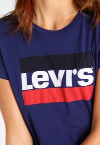Námořnická modrá bavlněná tričko s krátkým rukávem, na které je velké bílé logo "Levi’s" orámované červenou a černou barvou přes hrudník.