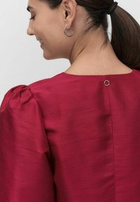 Rød, tekstureret bluse med puffede ærmer og en rund halsudskæring. Har en lille cirkulær knaplukning bagpå.