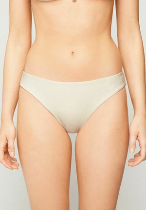 Femme portant un bas de bikini blanc uni, montrant la taille et le haut des jambes sur un fond neutre.