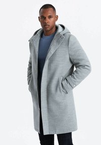 Abrigo gris con capucha, cierre de cremallera, bolsillos laterales y una textura suave. Diseño en capas sobre una camiseta azul acanalada.